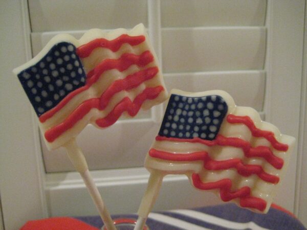 Two 12-White Chocolate American Flag Lollipops, featuring red, white, and blue colors on sticks, are displayed in front of white shutters.