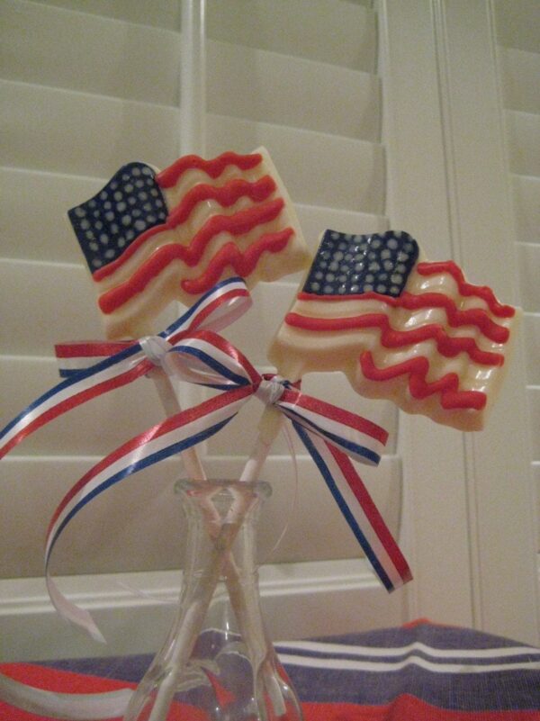 Two white chocolate American flag lollipops featuring red, white, and blue ribbons are displayed in a glass vase in front of a window with white shutters.
