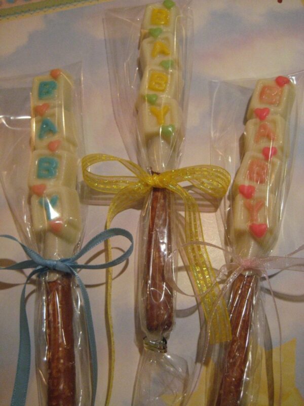 Three chocolate-covered pretzel rods wrapped in clear plastic, tied with blue, yellow, and pink ribbons. Each rod has colored letters spelling "BABY." Product: 24-Chocolate Covered Pretzel Rods-BABY Verse-Baby Shower Favors.