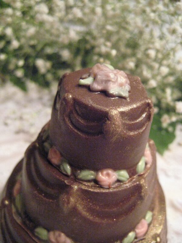 The 12-Elegant Solid Chocolate Wedding Cake Favor is a three-tiered chocolate cake, adorned with green and pink floral icing, complemented by a backdrop of white flowers.