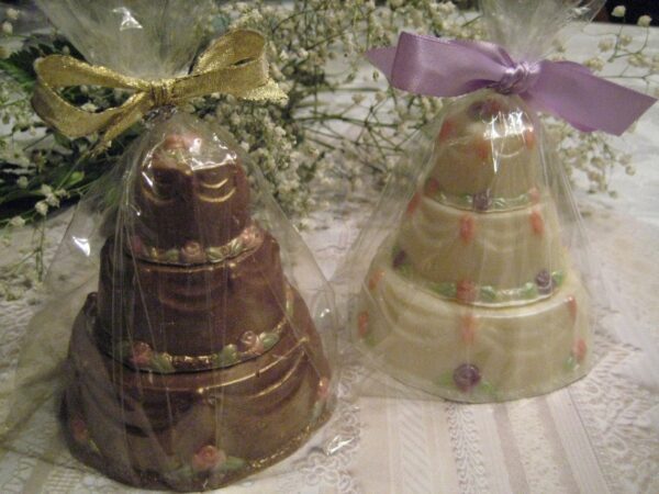 Two decorative 12-Elegant Solid Chocolate Wedding Cake Favors, tiered and wrapped in clear plastic, feature one with a gold ribbon and the other with lavender ribbon, adorned with delicate flowers atop a lace tablecloth.