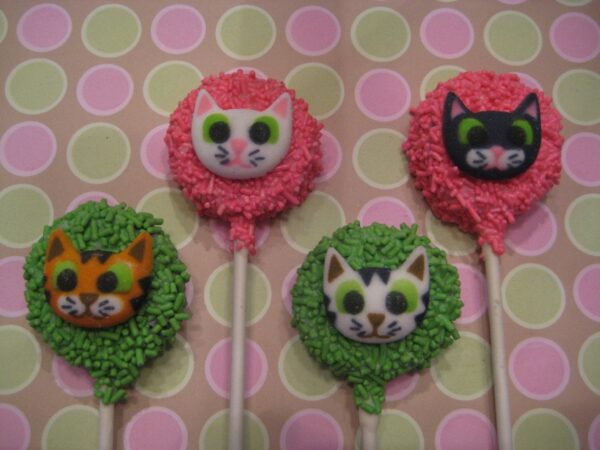 Four Oreo pops, each adorned with different cute kitty faces, colorful icing, and sprinkles. They are set against a pastel pink and green polka-dotted background.