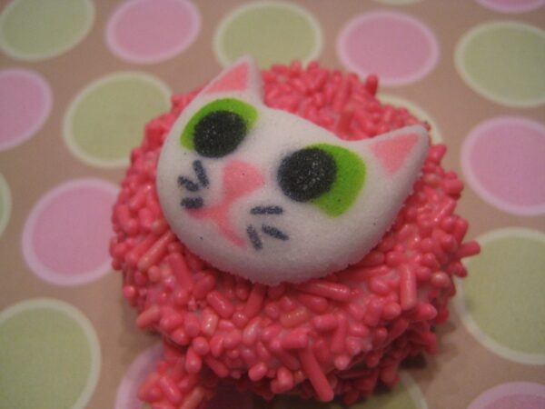 A pink sprinkled cupcake with a cute cat face decoration featuring green eyes and pink ears, set against a pastel circle background. Product: 12 Cute Kitty Faces On Chocolate Covered Oreo Pops.