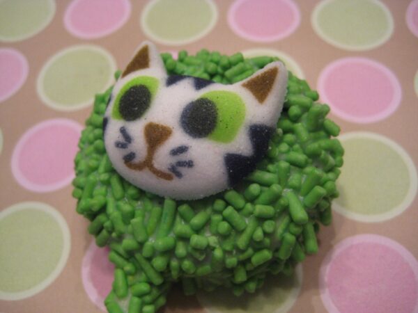 A chocolate-covered Oreo pop featuring a cute kitty face with green sprinkles sits against a pink and green polka-dotted background.