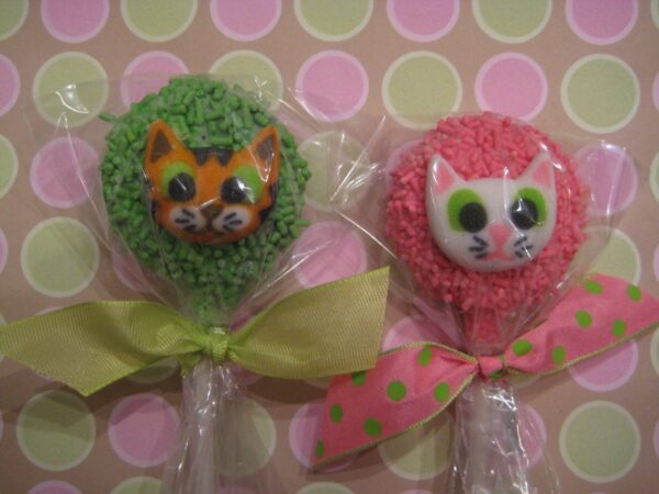 Two cat-themed Oreo pops wrapped in cellophane; one green with an orange cat, one pink with a white cat, both adorned with polka dot ribbons on a polka dot background from "12 Cute Kitty Faces On Chocolate Covered Oreo Pops".