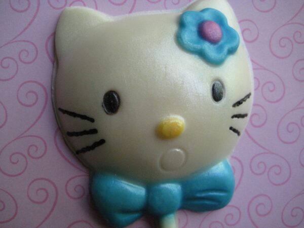 A 12-pack of Hello Kitty-shaped white chocolate lollipops with a blue bow and flower against a swirled pink background.
