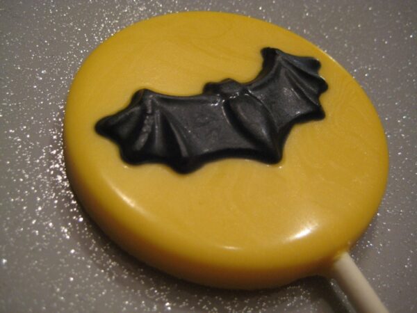 A round yellow lollipop showcases a black bat design on a glittery gray surface, from the 24-Chocolate Round Bat Lollipops collection for party or Halloween favors.