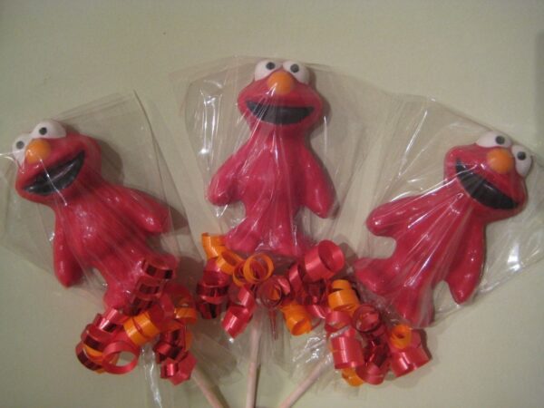Three Elmo lollipops with wide eyes and smiles, wrapped in clear plastic and adorned with red and orange curly ribbons.