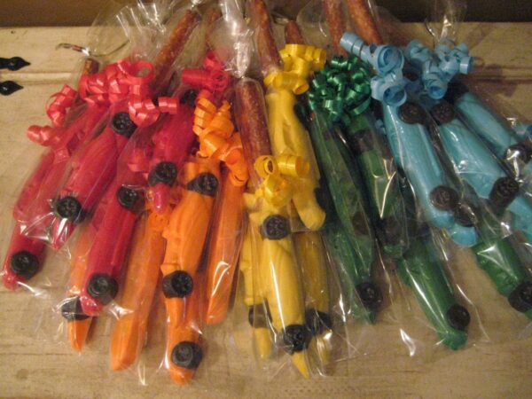 A wooden surface displays colorful bundles of 24-Race Car Chocolate Covered Pretzel Rods, each wrapped with curly ribbons and round embellishments in red, orange, yellow, green, and blue.