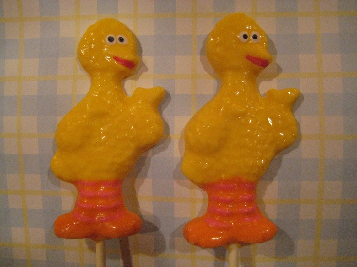 Two yellow Big Bird chocolate lollipops with orange feet and pink bases from the 12-pack set, ideal for children's birthday party favors, against a checked background.