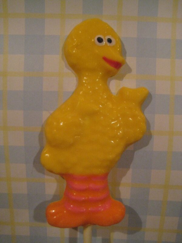 A Big Bird chocolate lollipop shaped like a yellow bird with red feet and big eyes, perfect for children's birthday parties, stands against a checkered background.