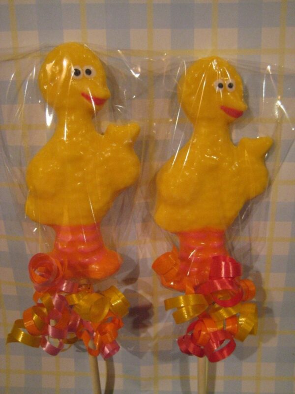 Two yellow, duck-shaped chocolate lollipops from a set of 12 with orange feet and colorful ribbons, wrapped in clear plastic against a checkered background.