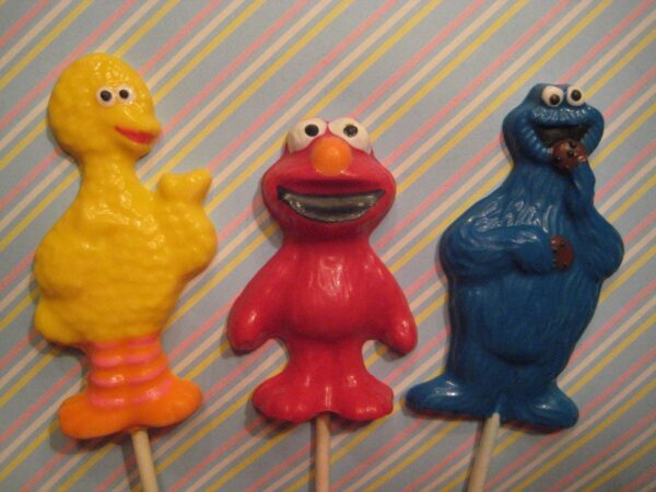 Three lollipops in blue monster, yellow bird, and red character shapes against a striped background; includes 12-chocolate Blue Monster Lollipop Favors for a children's birthday party.