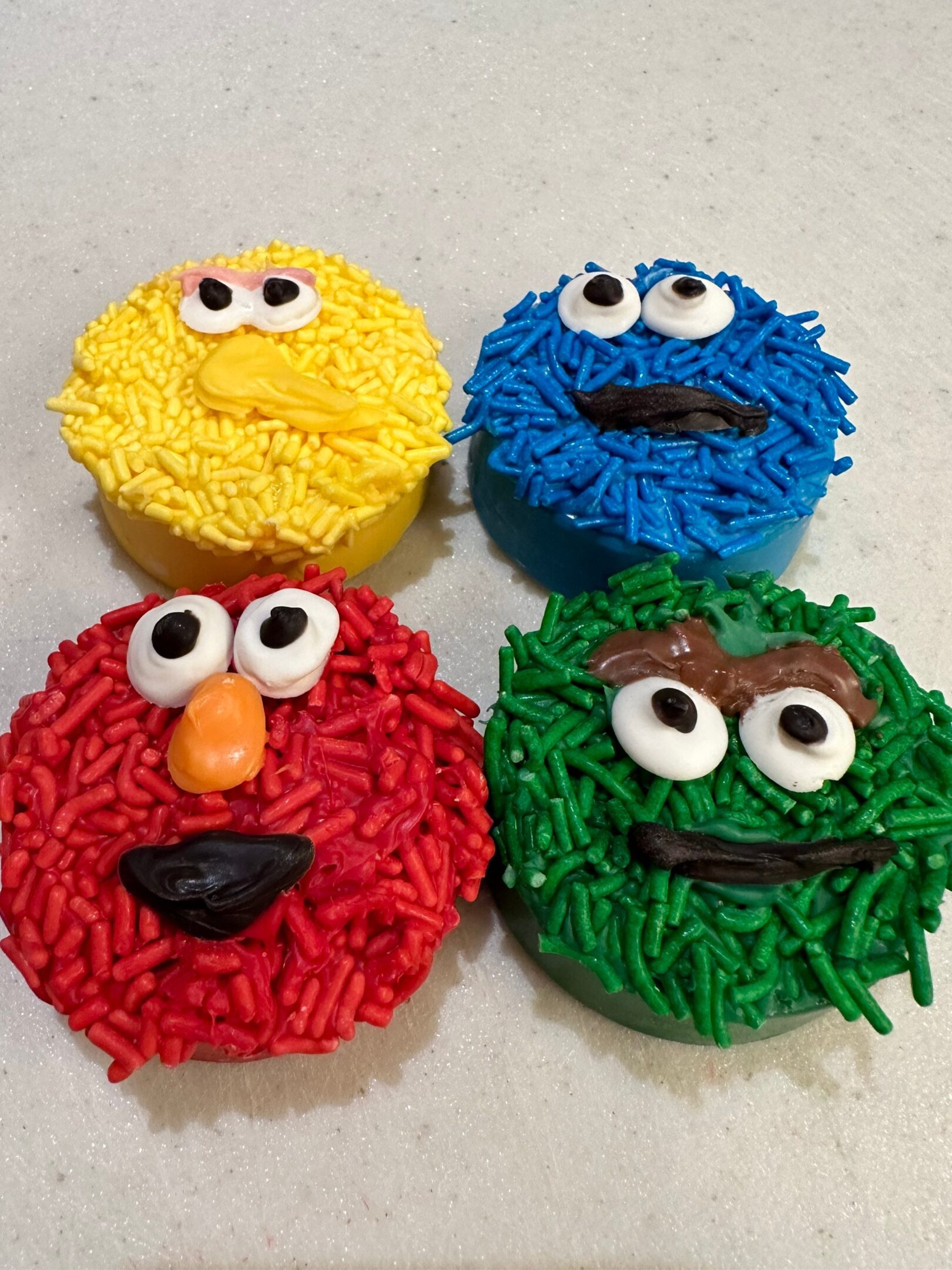 A set of 24-Sesame Monster Chocolate Covered Oreos meticulously decorated with colorful icing and candy eyes to resemble beloved children’s show characters, showcasing vibrant red, yellow, blue, and green faces.