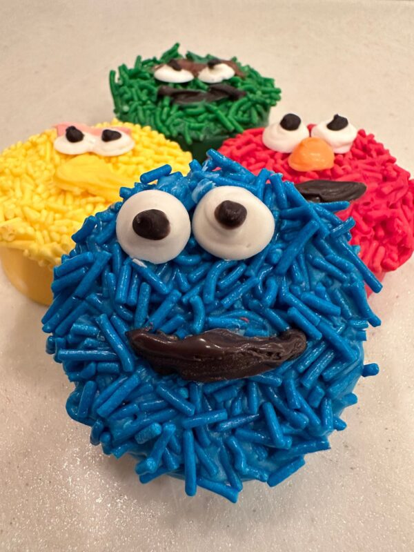Four 24-Sesame Monster Chocolate Covered Oreos feature playful characters with blue, red, yellow, and green icing faces and edible eyes on a white surface.