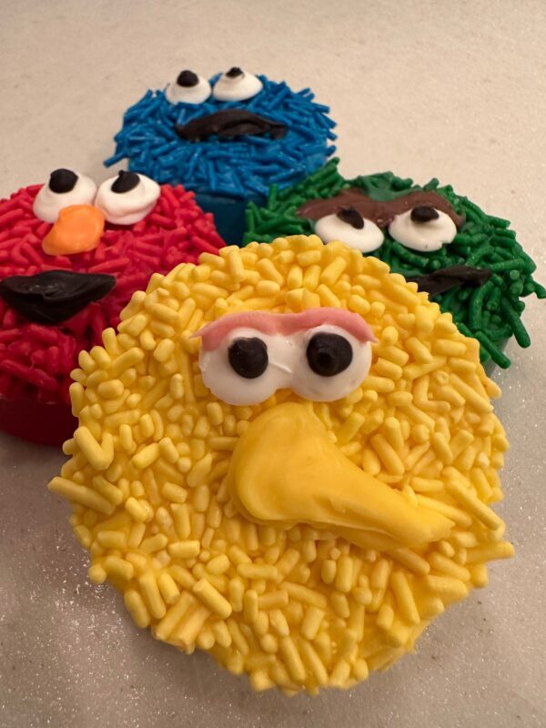 Four Sesame Monster Chocolate Covered Oreos decorated with vibrant frosting and sprinkles to resemble puppet characters, complete with distinct eyes and features.