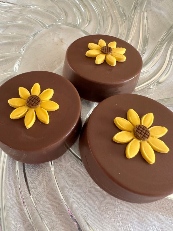 Three Sunflower Milk Chocolate Covered Oreos with yellow sunflower designs are artfully arranged on a clear, textured plate.
