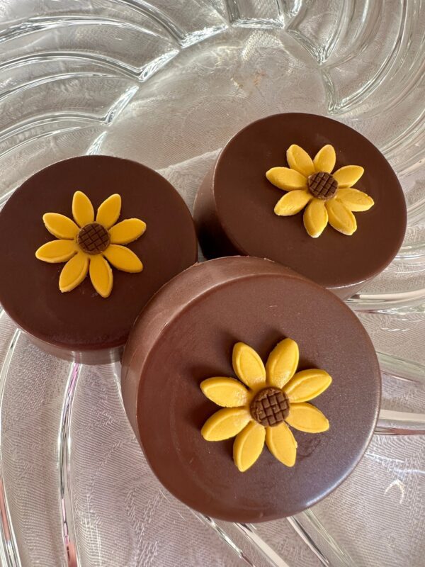 Three 12-Sunflower Milk Chocolate Covered Oreos on a glass plate, each topped with a sunflower decoration made from yellow and brown icing.