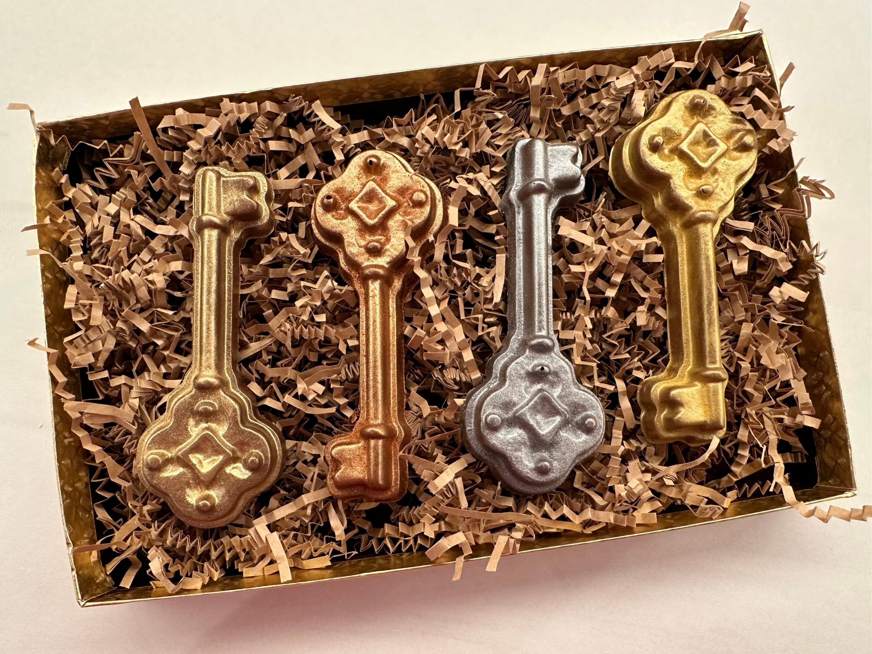 The 4-Milk Chocolate Keys Gift Box showcases keys in two gold, one bronze, and one silver color displayed on crinkle-cut paper.