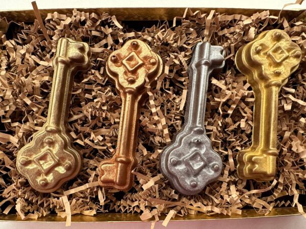 The 4-Milk Chocolate Keys Gift Box includes four metallic key-shaped chocolates—two gold, one bronze, and one silver—arranged on shredded paper in a rectangular box.
