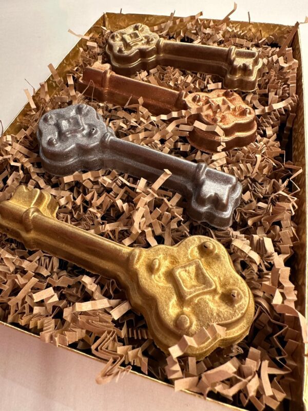 A decorative box containing four milk chocolate keys—two gold, one bronze, and one silver—placed on shredded paper.