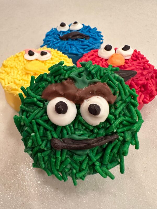 Four colorful, icing-decorated chocolate-covered Oreos with candy eyes, resembling muppet characters, are arranged on a white surface.