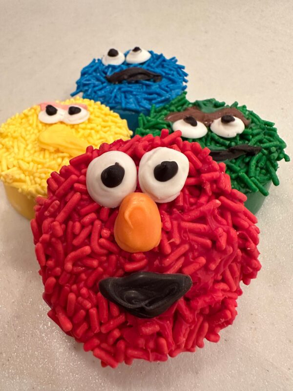 Four Sesame Monster chocolate-covered Oreos decorated like colorful, furry puppet characters with expressive faces using red, yellow, blue, and green icing and candy eyes.