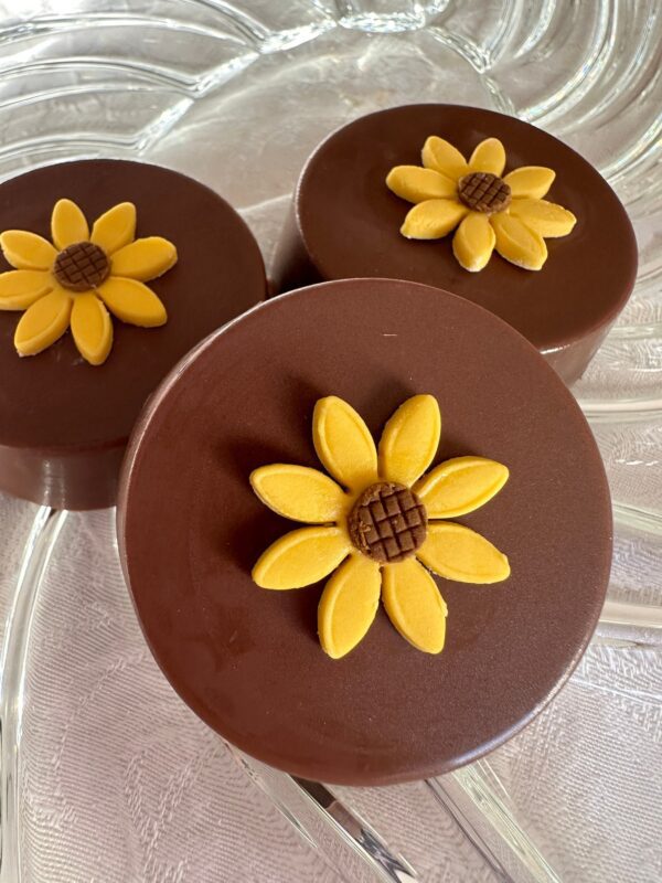 Three milk chocolate-covered Oreos, each topped with a yellow sunflower decoration, are displayed on a clear glass plate.