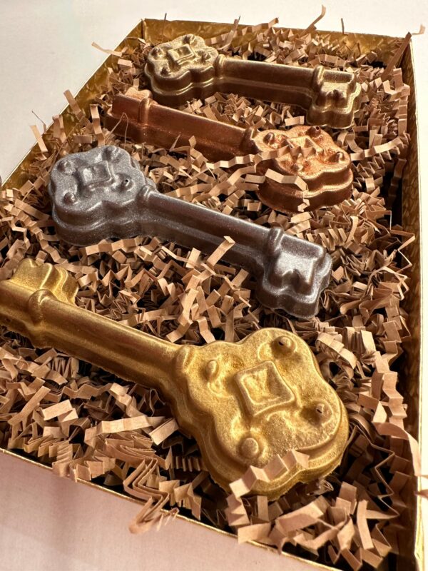 The 4-Milk Chocolate Keys Gift Box includes two gold, one bronze, and one silver ornate key-shaped chocolates presented in a box with crinkle paper.