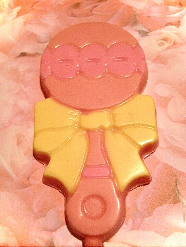 A pink and yellow lollipop shaped like a rattle with a bow, set against a floral pink background.