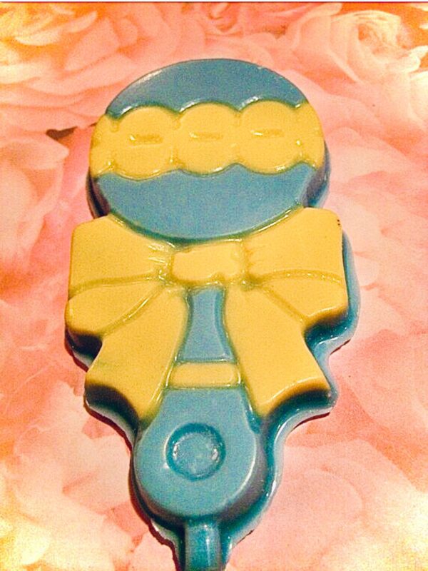 A blue and yellow chocolate baby rattle lollipop topped with a bow, set against a pink floral background.