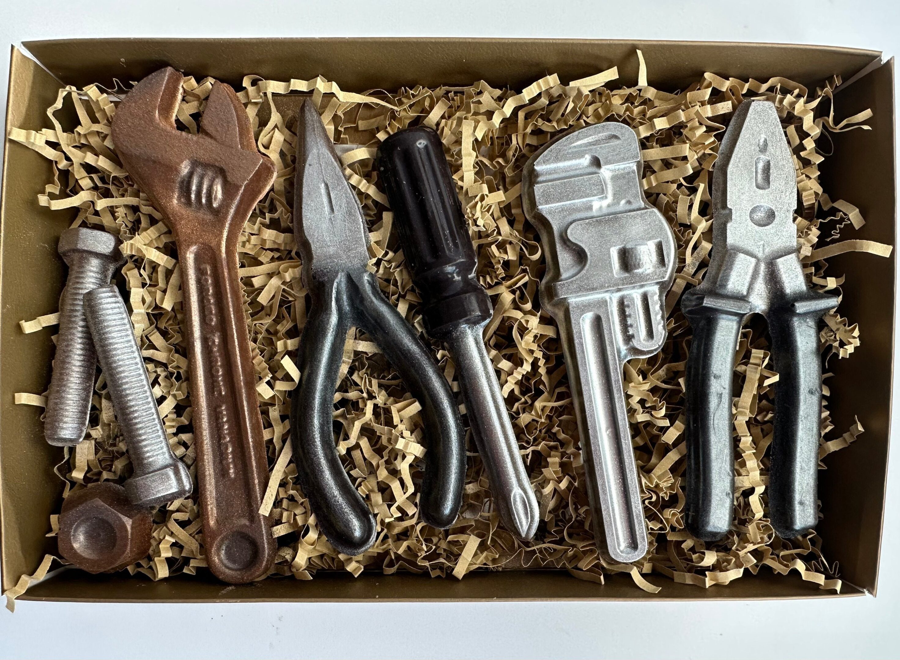 The 8 Piece Chocolate Tool Gift Set features tool-shaped chocolates, including a wrench, pliers, screwdriver, pipe wrench, and bolt cutter, elegantly presented on shredded packing material.