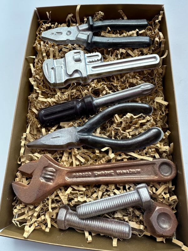 The 8 Piece Chocolate Tool Gift Set includes a variety of chocolate tools like a wrench, pliers, screwdriver, and bolts, beautifully arranged on shredded paper in a gift box.