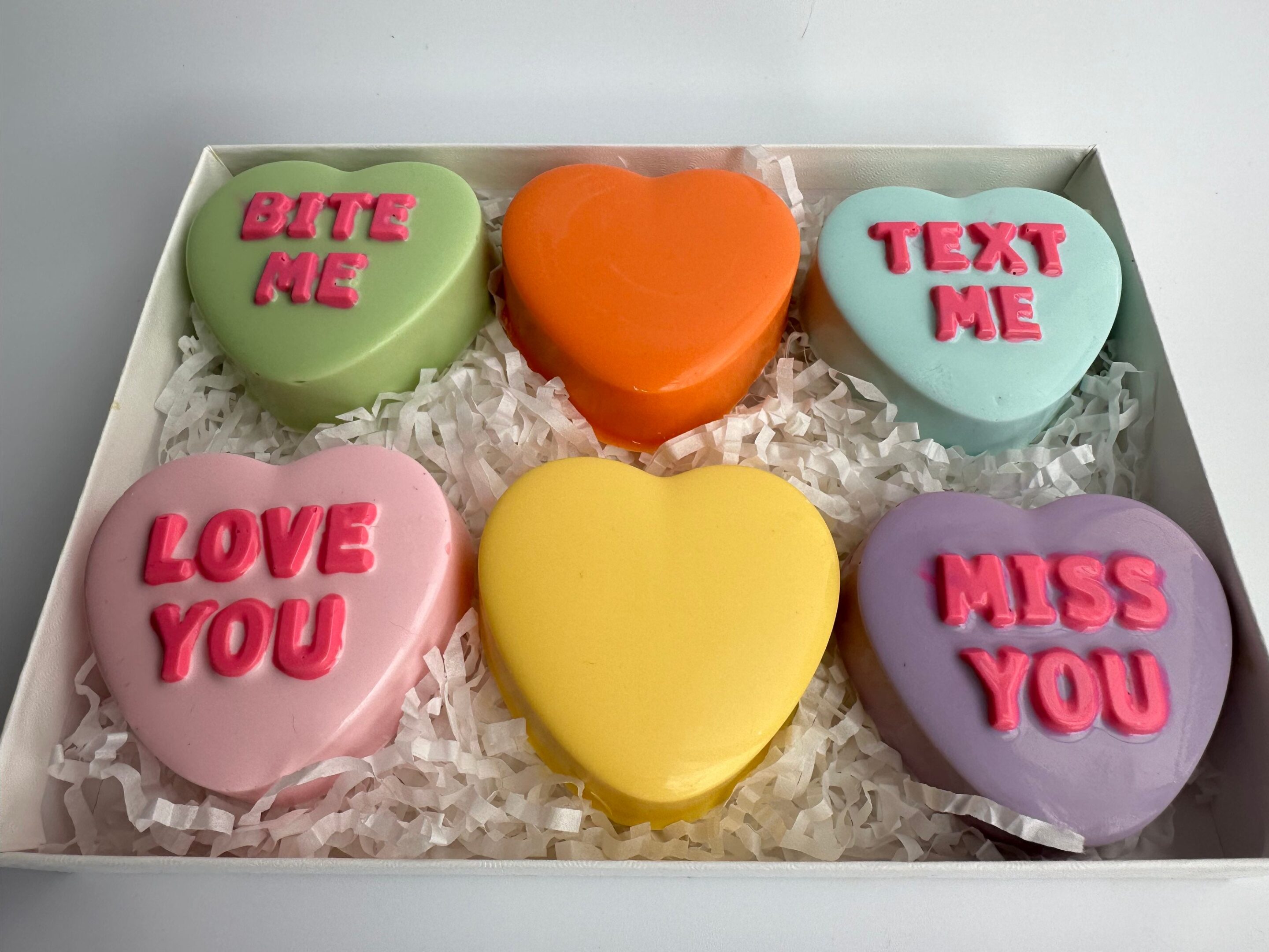 Gift box of six heart-shaped Chocolate Covered Oreos with pastel icing, featuring messages like "Bite Me," "Text Me," "Love You," "Miss You" in pink frosting, on shredded paper.