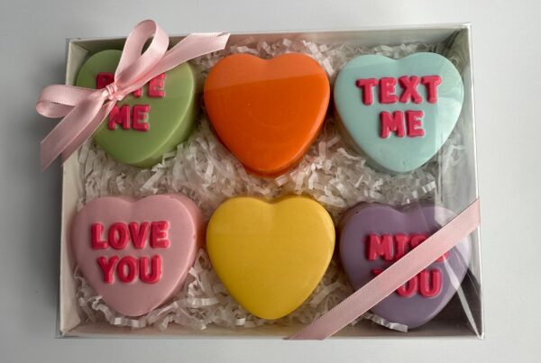 Gift box of six heart-shaped chocolate-covered Oreos with colorful icing reads: "Kiss Me," "Text Me," "Love You," "Miss You," plus two plain hearts, nestled in white shredded paper.