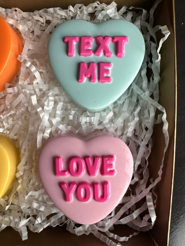 The 6 Conversation Heart Chocolate Covered Oreos Gift Box features heart-shaped chocolates, with messages like "TEXT ME" on blue and "LOVE YOU" on pink, nestled in white shredded paper.