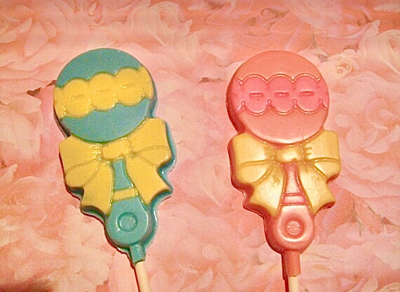 Two baby rattle-shaped lollipops, one blue and one pink with yellow bows, from the "12-Chocolate Baby Rattle Lollipop For Baby Shower Favors/Baby Gift" collection are placed on a pink floral background.