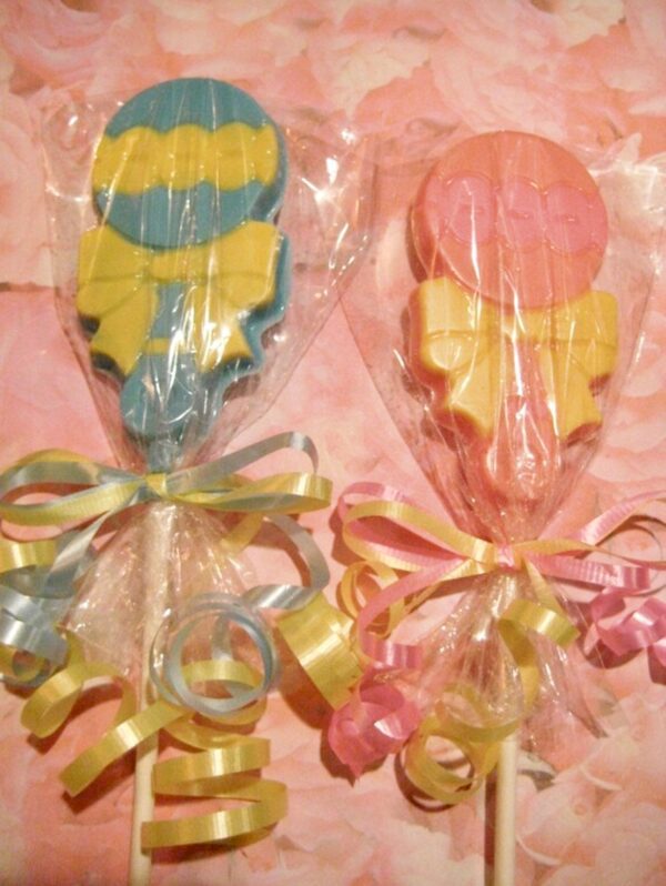 Two 12-Chocolate Baby Rattle Lollipops for baby showers, wrapped in clear plastic. One is blue with a yellow ribbon, the other pink with a yellow ribbon, set against a pink background.
