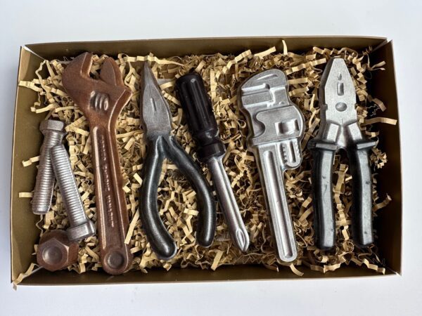 The 8 Piece Chocolate Tool Gift Set features chocolate replicas of tools like a wrench, pliers, screwdriver, and nuts, all nestled on crinkled paper shreds.