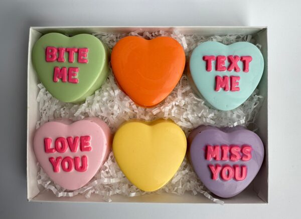A box of six chocolate-covered Oreos in heart shapes, featuring "Bite Me," "Text Me," "Love You," "Miss You" messages, plus one blank orange heart. Product: 6 Conversation Heart Chocolate Covered Oreos Gift Box.