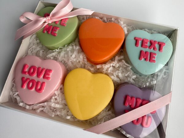 A gift box containing six heart-shaped chocolate-covered Oreos in assorted colors, embossed with "LOVE ME," "TEXT ME," "LOVE YOU," and "MISS YOU," and adorned with a pink ribbon.
