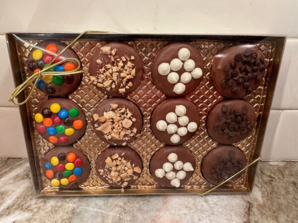 A 12-chocolate covered Oreo cookies gift box, topped with colorful candies, chopped nuts, mini marshmallows, and chocolate chips, is arranged in a gold-lined tray with a gold ribbon.