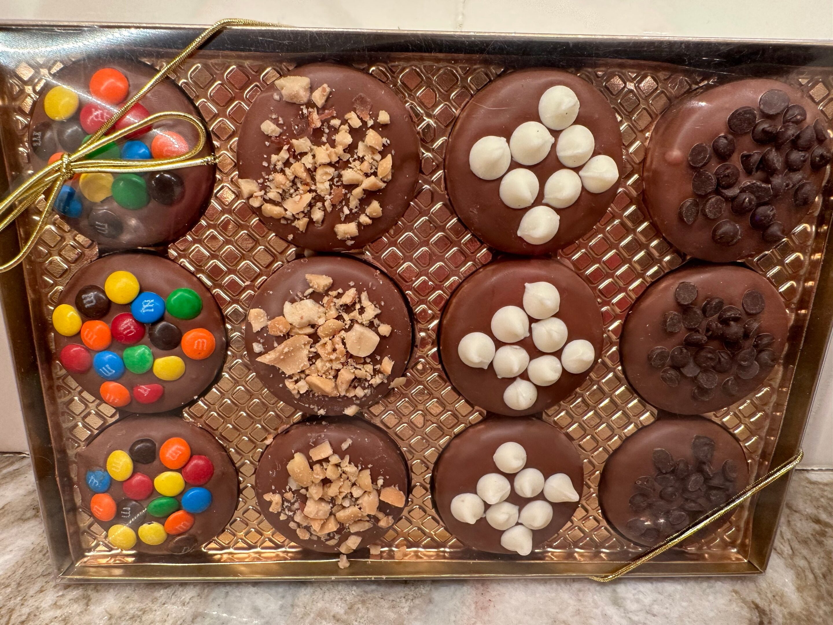 A 12-piece gift box of chocolate-covered Oreo cookies topped with M&Ms, crushed nuts, marshmallows, and chocolate chips on a gold lattice tray.