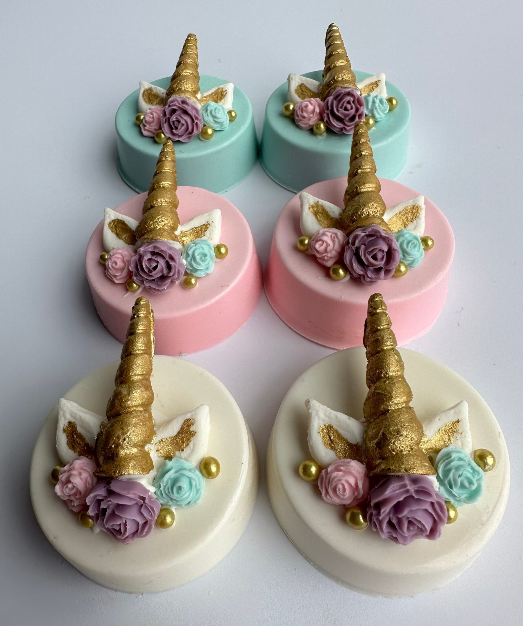 Six unicorn-themed chocolate-covered Oreos with golden horns, adorned with roses, pastel colors, and edible pearls.