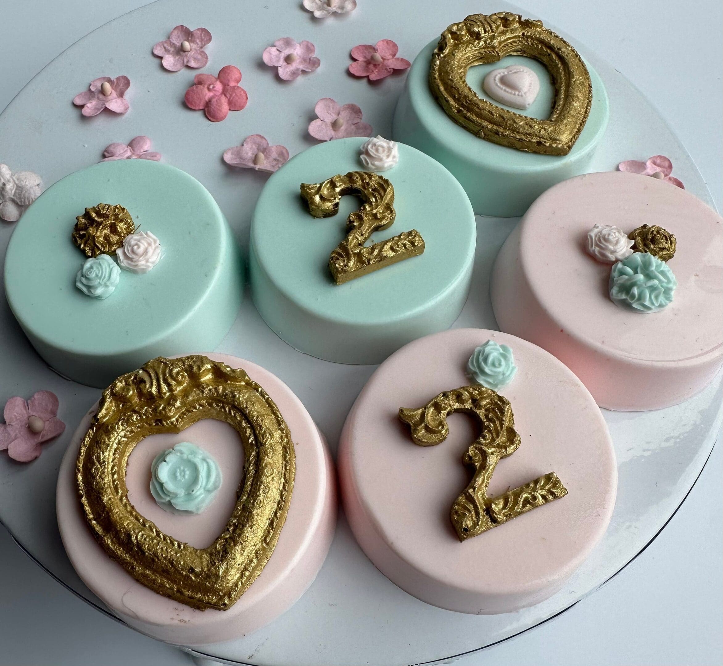Five pastel cakes with ornate gold numbers are surrounded by small pink and white flower decorations, much like the 12-Gold Frame Chocolate Covered Oreos for Girl or Boy Birthday with Age.