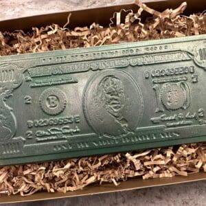 A rectangular green bar resembling a U.S. hundred-dollar bill is nestled in crinkled paper within the "Chocolate 100 Dollar Bill Gift Box.