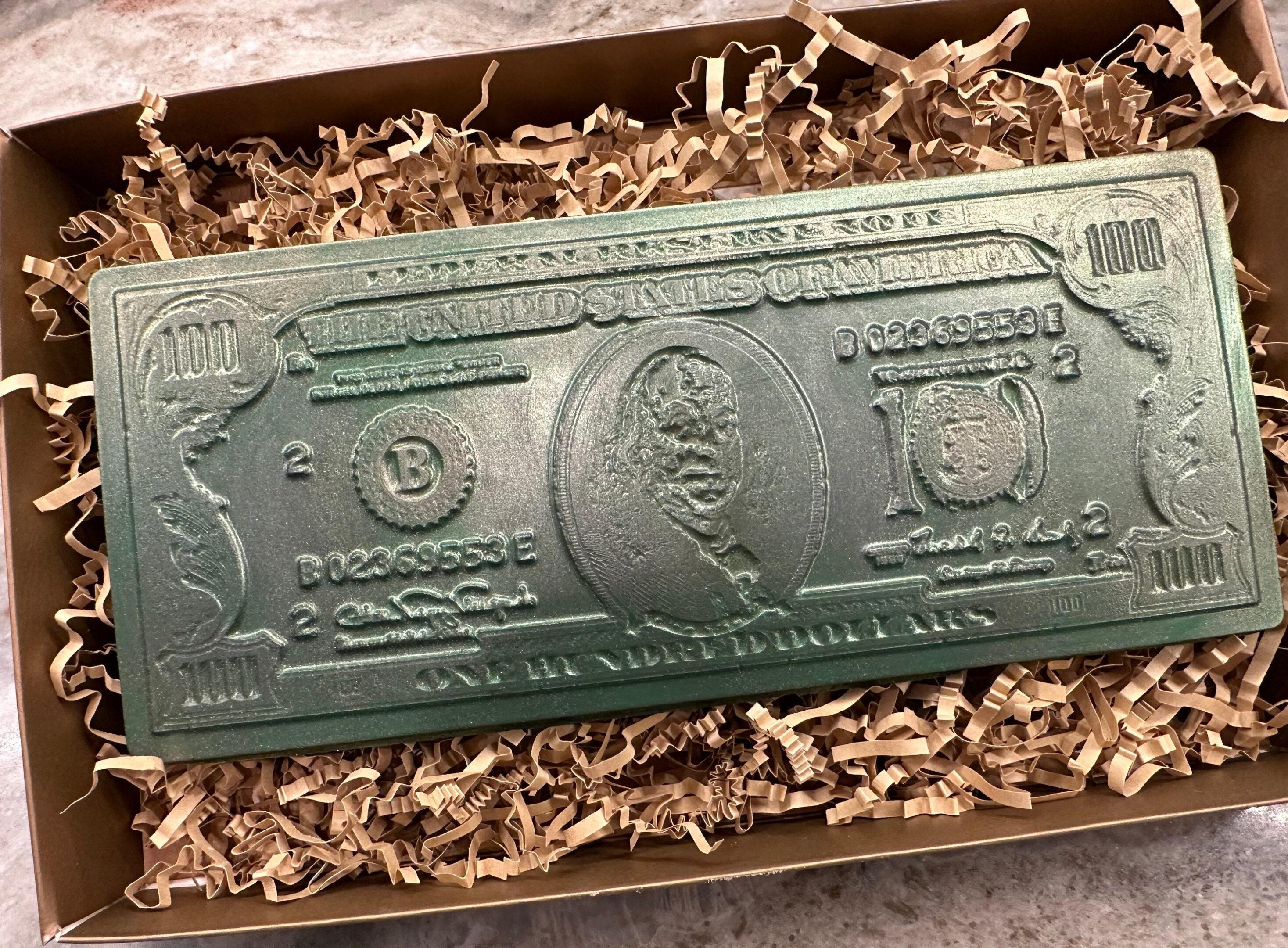 A rectangular green bar resembling a U.S. hundred-dollar bill is nestled in crinkled paper within the "Chocolate 100 Dollar Bill Gift Box.