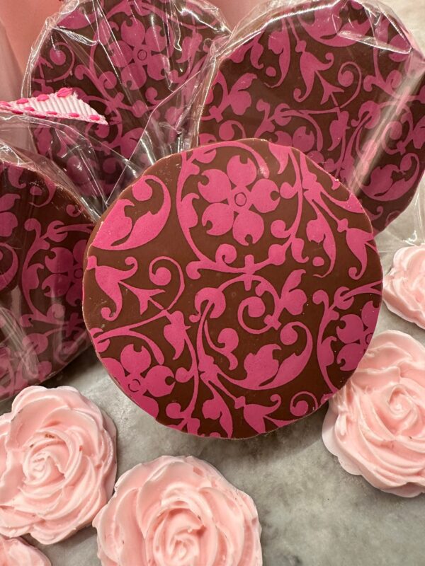 Displayed upright on a table, the 12-Pink Floral Scroll Chocolate Covered Oreo Gift Box Set includes pink and brown patterned chocolate discs alongside decorative pink rose-shaped candies.