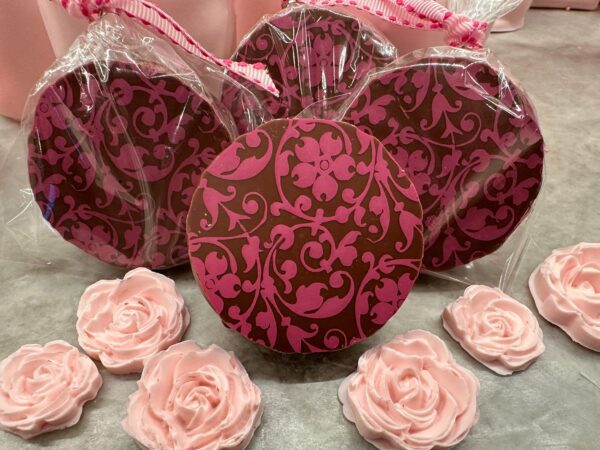 A 12-piece gift box of pink and brown floral scroll chocolate-covered Oreos, wrapped in plastic, elegantly displayed with pink rose-shaped decorations on a gray surface. Perfect for weddings, birthdays, parties, or baby showers.