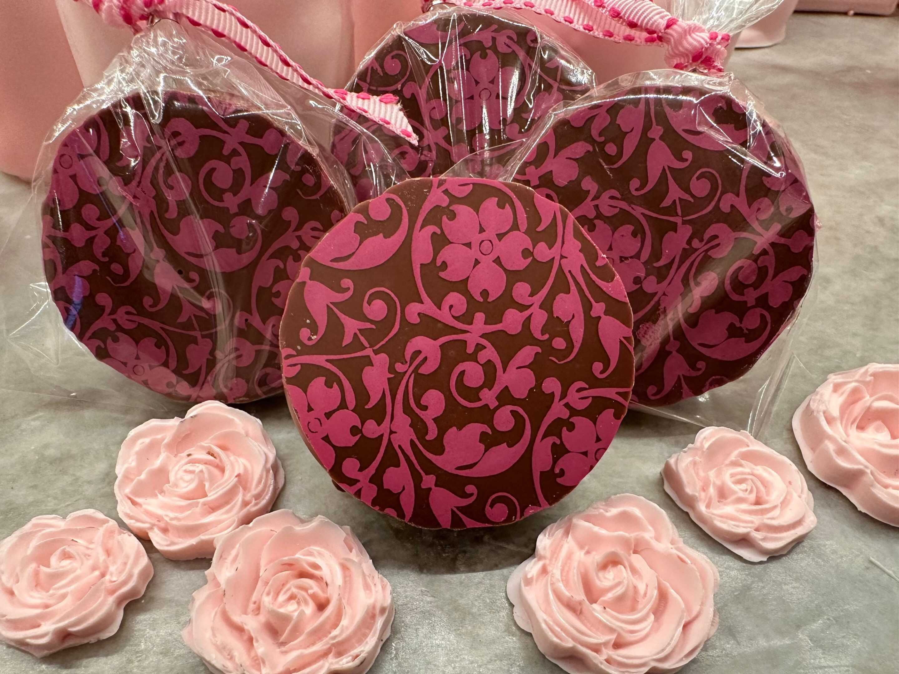 A 12-piece gift box of pink and brown floral scroll chocolate-covered Oreos, wrapped in plastic, elegantly displayed with pink rose-shaped decorations on a gray surface. Perfect for weddings, birthdays, parties, or baby showers.
